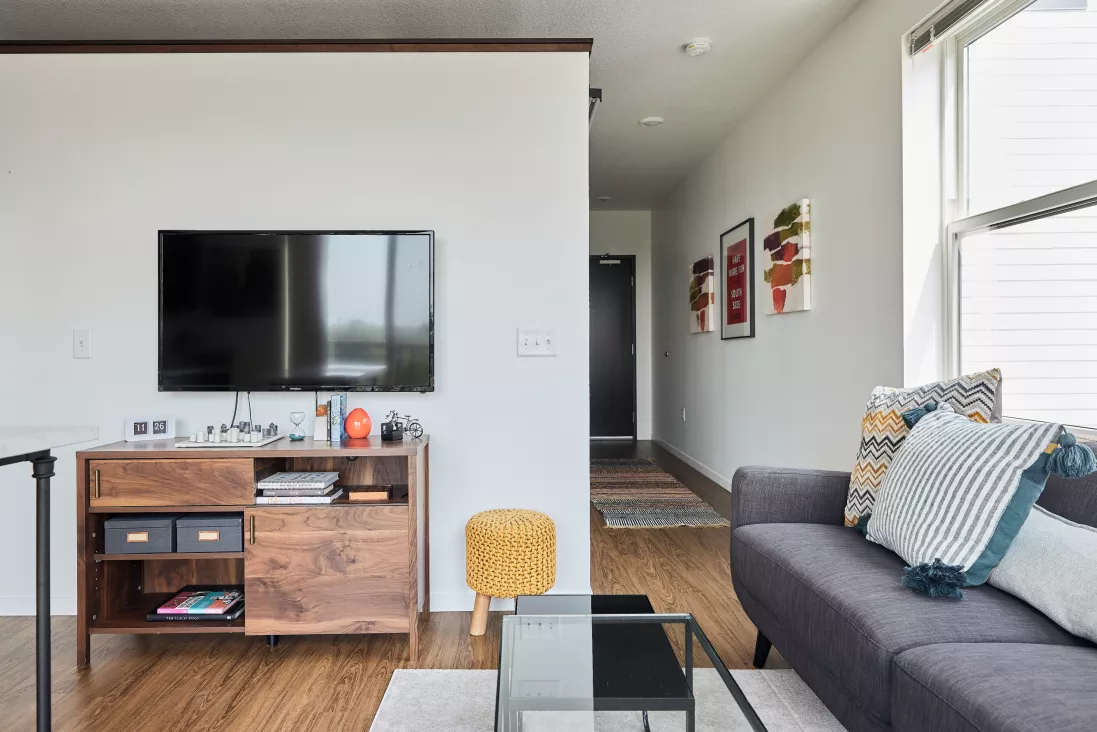 Modern living room with a mounted TV, wooden media console, and gray sofa with pillows. Hallway with artwork in background.