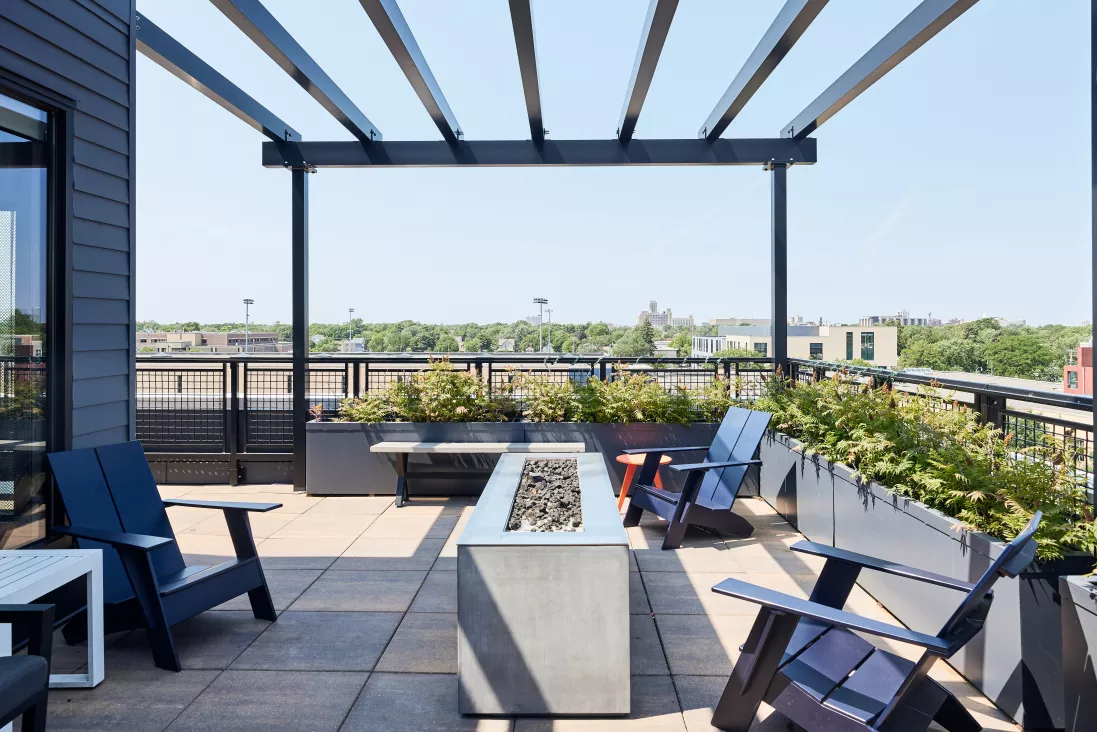 Outdoor rooftop terrace with lounge chairs, a linear fire pit, and lush green plants. Clear sky above city.